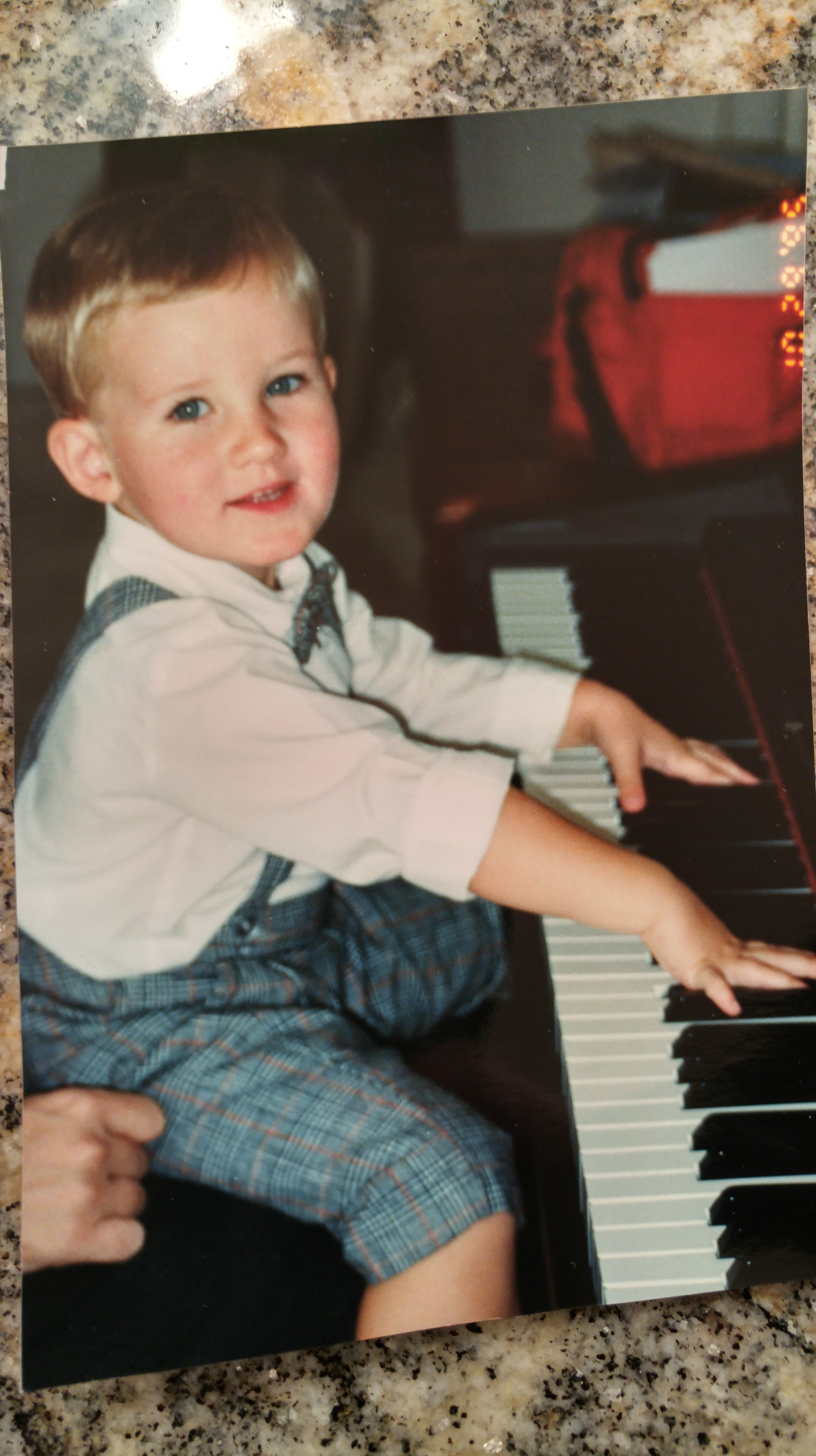 A young boy with short blonde hair, dressed in a white shirt and plaid shorts, sitting at a piano and smiling while playing.